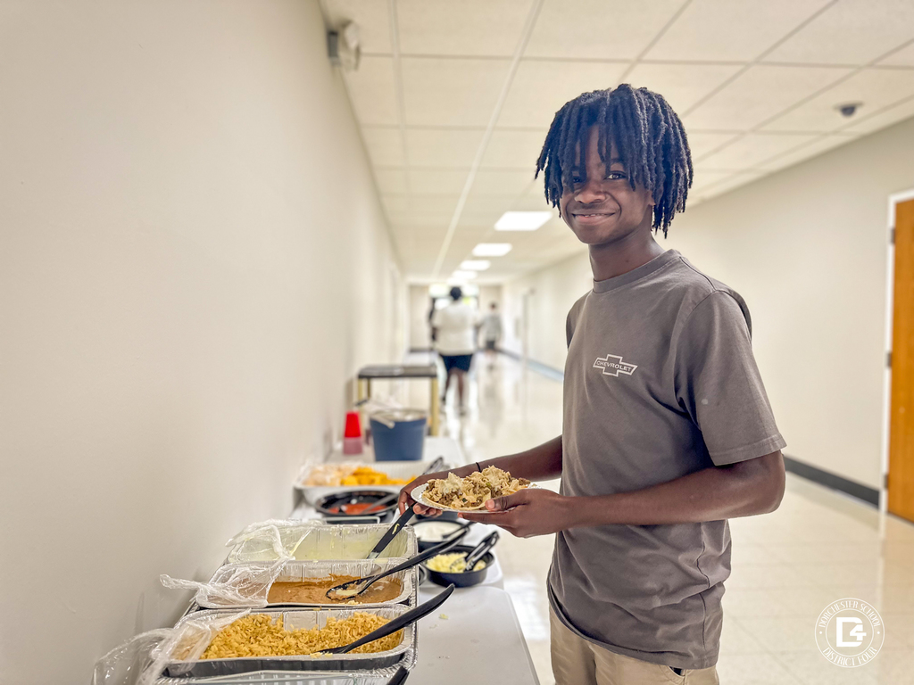 A young man wearing a gray Chevrolet shirt smiles while serving himself lunch from a buffet line set up in a school hallway, celebrating the end of the Summer Work Crew program.