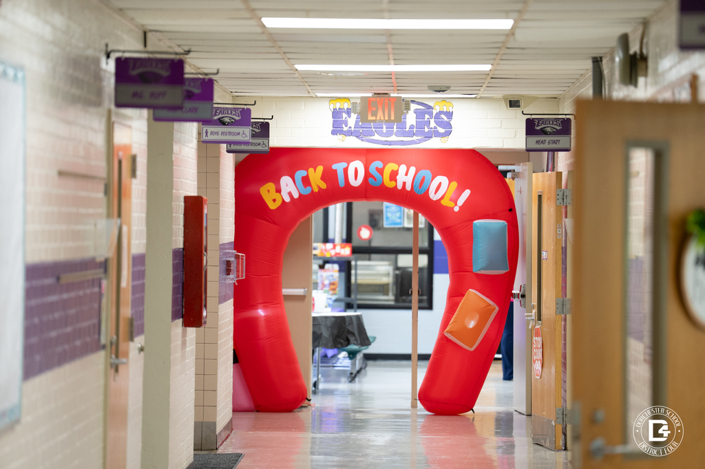 A brightly colored, inflatable red archway with the words "Back to School!" stands in a school hallway. The arch is decorated with blue, orange, and yellow book-shaped accents. Purple signage above and around the hallway identifies classrooms and restrooms, and a sign labeled “Eagles” is visible above the arch. The setting is cheerful and welcoming, celebrating the return to school.