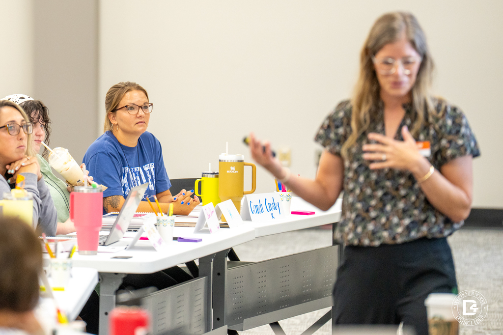 An instructor stands in front of the training room speaking, gesturing with one hand. Teachers seated at the tables are focused on the presentation. Name tags and large drink tumblers are on the tables.