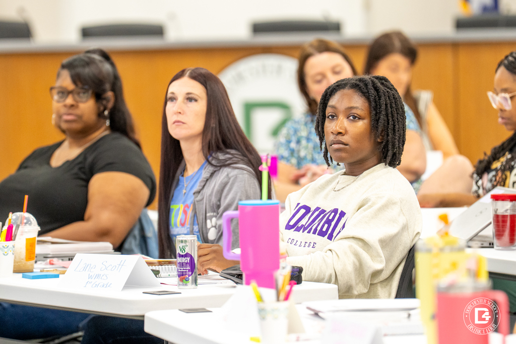 Several teachers at the front row of a professional development session, one wearing a Columbia College sweatshirt and another with a can of V8 Energy in front of her. Name tags and supplies are on the tables.