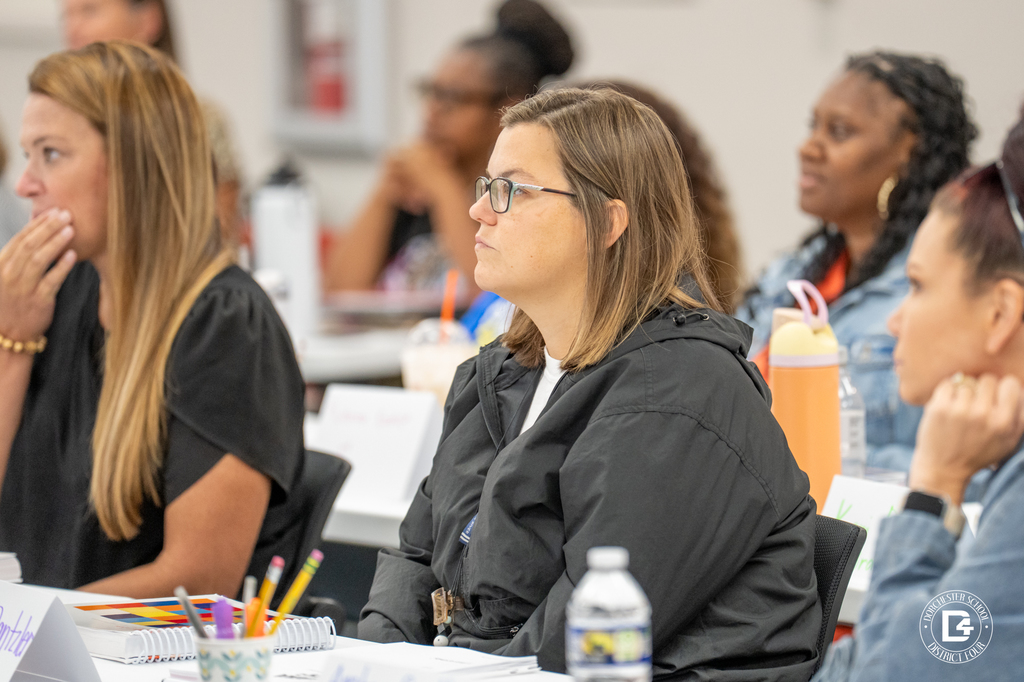 Close-up of a group of teachers seated at a training session, with one teacher in a black jacket attentively listening. Colorful notebooks and drinks are on the table.