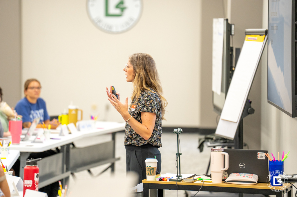 A facilitator stands beside a whiteboard and large screen, leading the training session. The Dorchester School District Four logo is visible behind her. A laptop, drink, and supplies are on a side table.