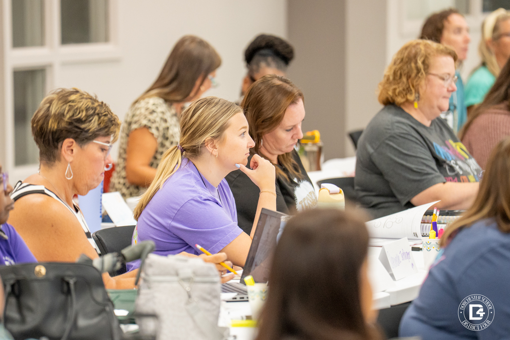 Teachers listening and taking notes during a training session. One teacher in a purple shirt is leaning forward with her hand on her chin. Training materials are open in front of the group.