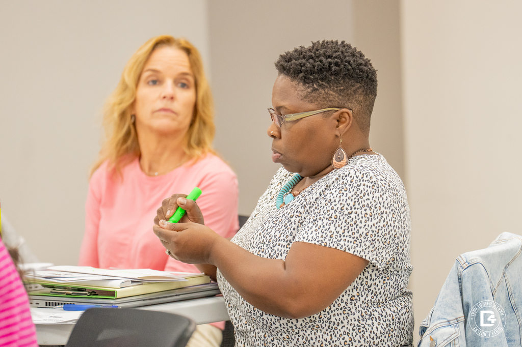 Two women seated at a training table, one focusing on a green highlighter marker while the other looks forward attentively. Binders and folders are spread out on the table in front of them.