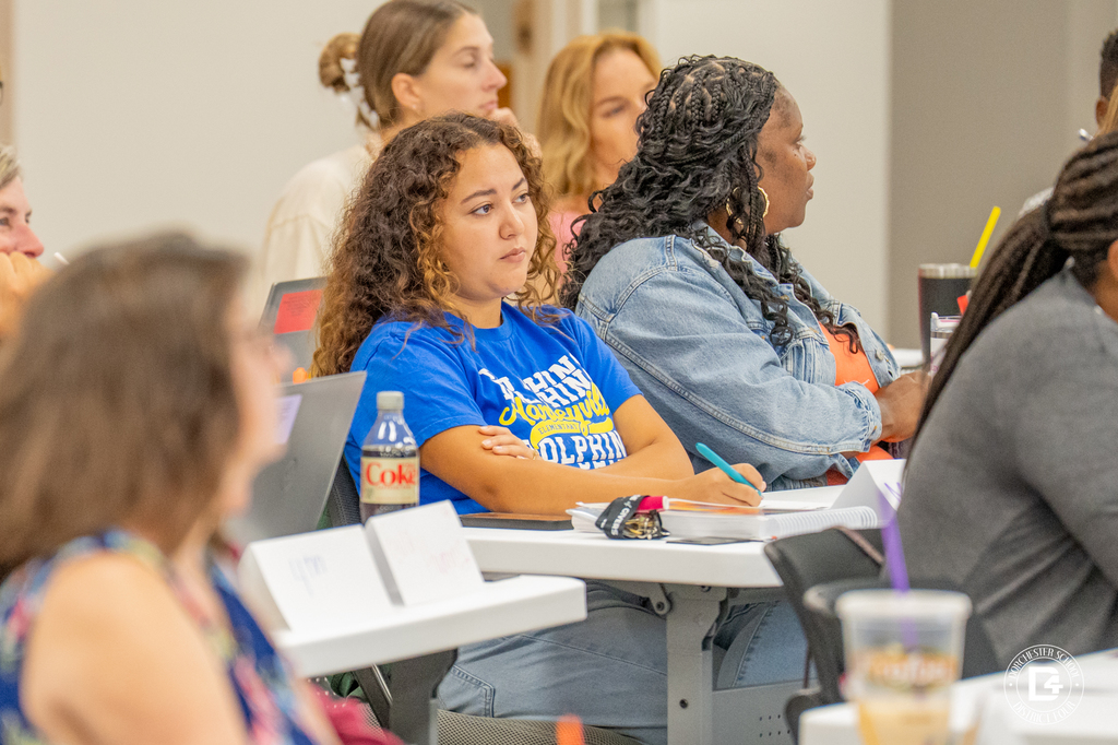 A teacher in a blue HES shirt takes notes during the session, sitting next to a colleague in a denim jacket. A Diet Coke bottle and notebook are on the table in front of her.