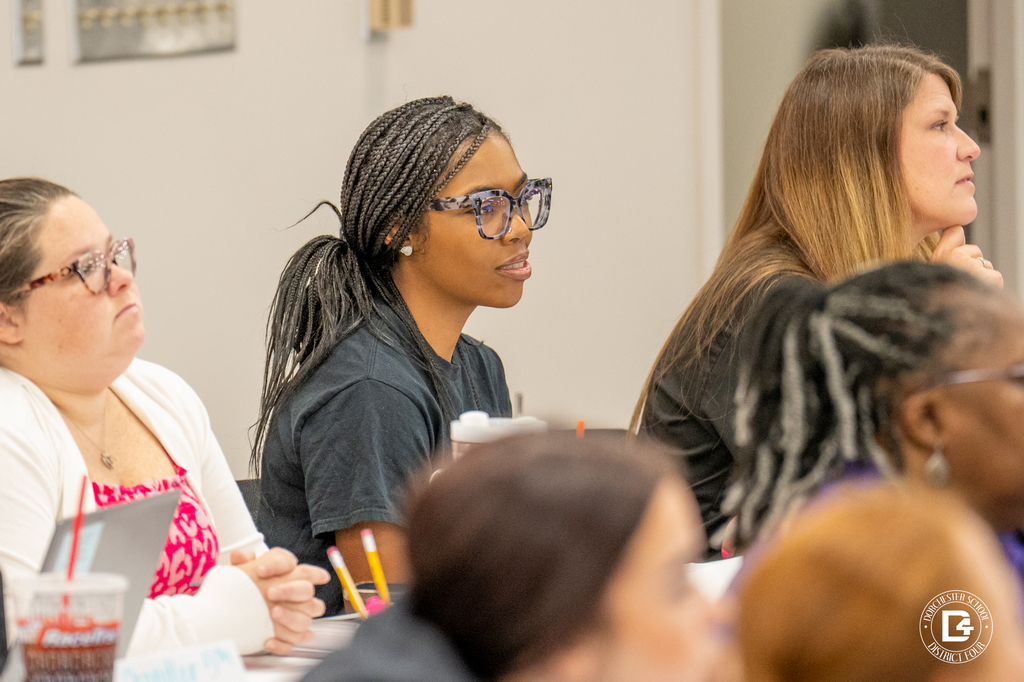 Close-up of teachers engaged in a professional development session. A woman in a black t-shirt with braided hair and large glasses listens attentively among a diverse group of colleagues.