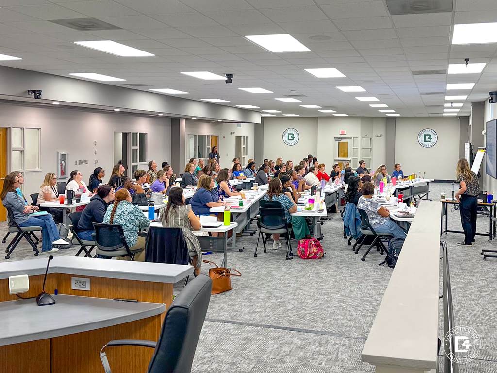 Wide shot of a large group of elementary teachers seated at tables in a district training room, listening to a speaker at the front. The room is well-lit with Dorchester School District Four logos on the walls.