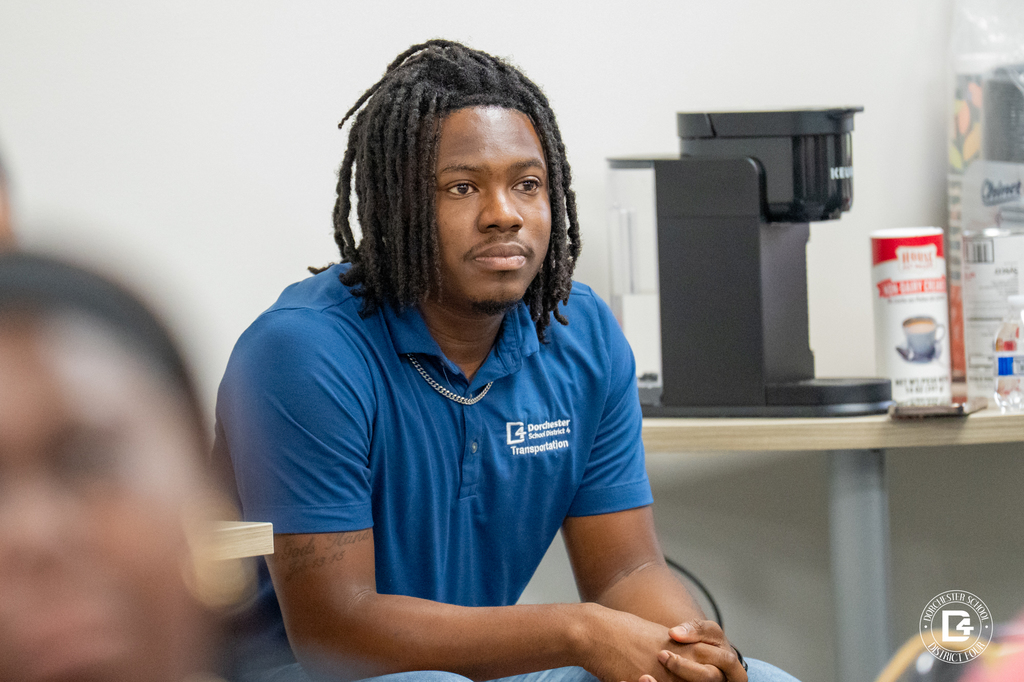 A young man in a blue Dorchester School District Four Transportation shirt sits attentively during the session, with a coffee maker in the background.
