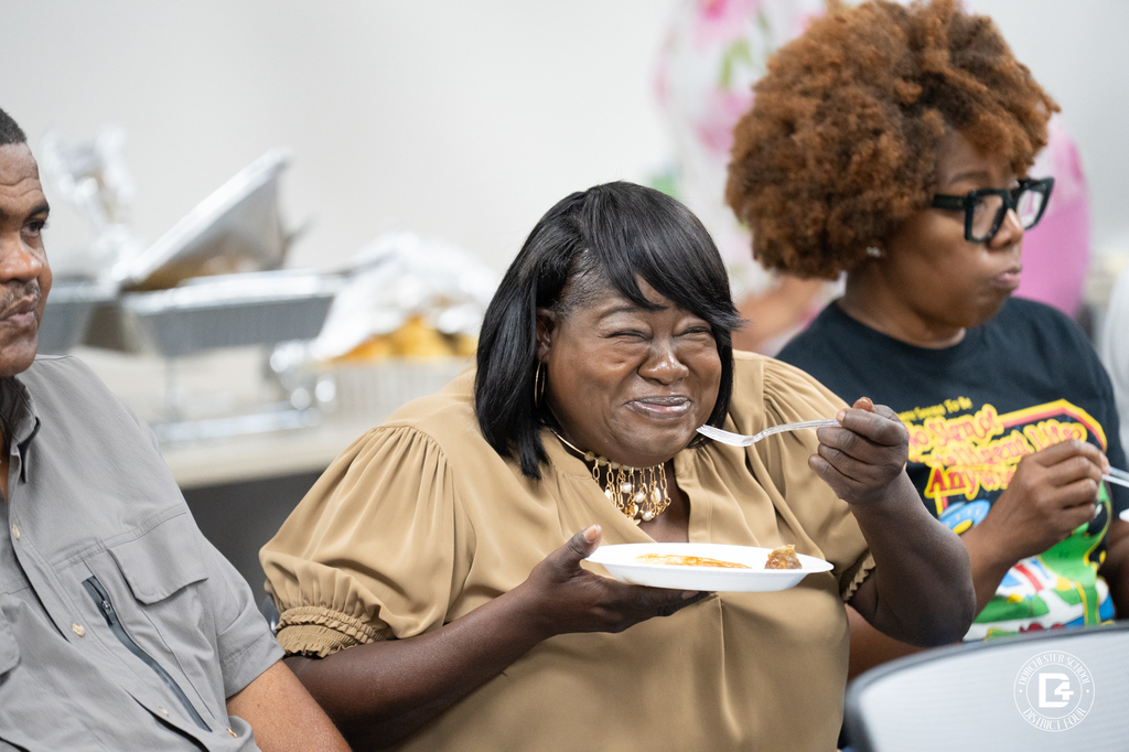 A woman in a tan top smiles joyfully while eating breakfast, seated beside other drivers during the meal portion of the event.