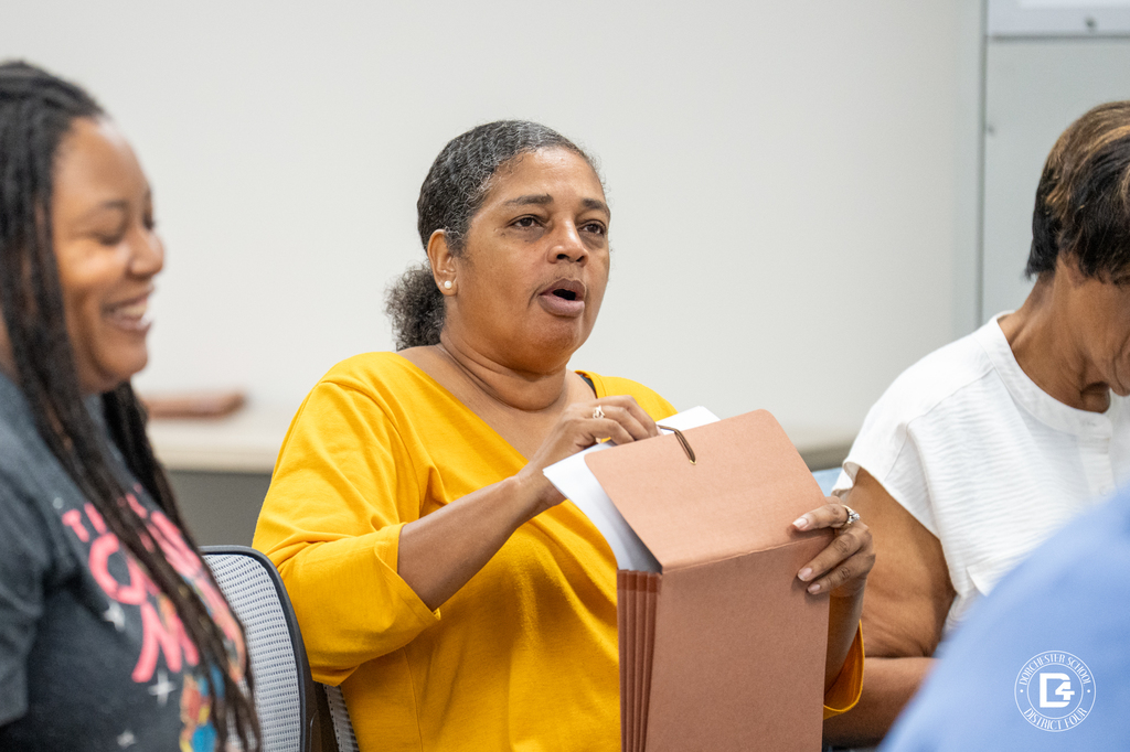 A woman in a yellow shirt opens a brown folder during the session, seated among other participants.