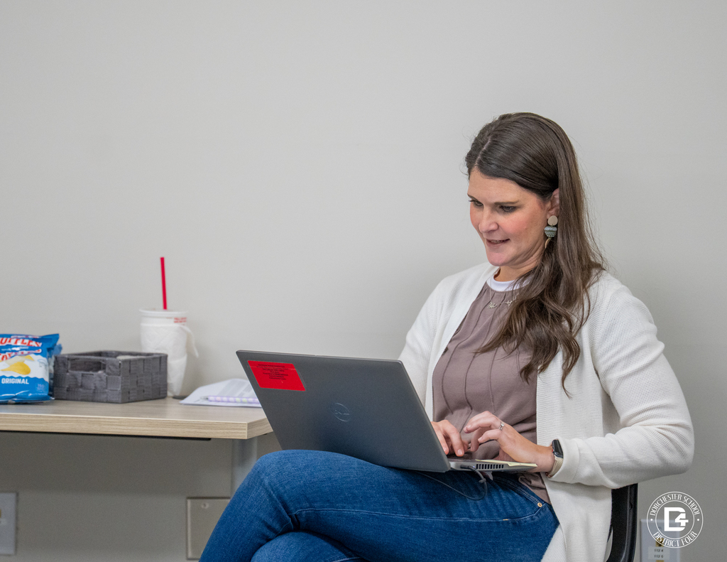 A female educator in a white cardigan sits to the side of the room, typing on her laptop with a drink and snack on the table beside her.