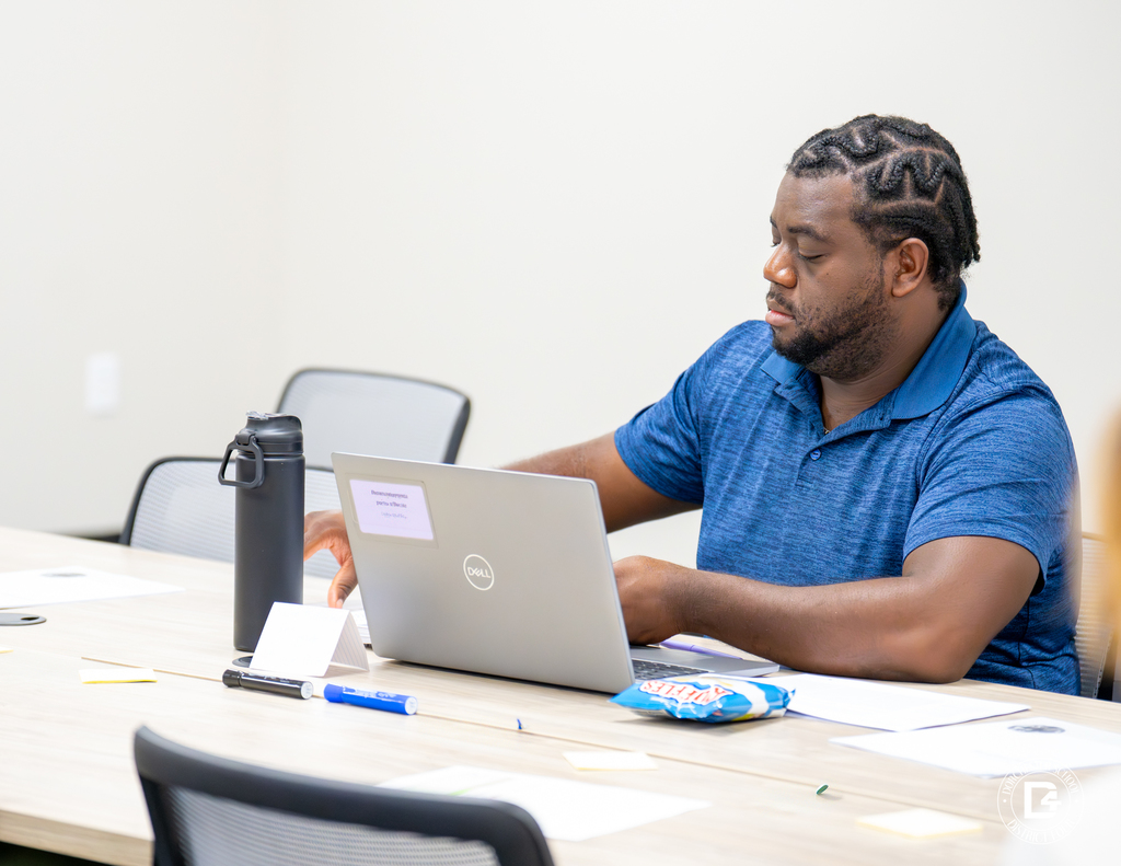 A male educator with braided hair works on a laptop during training, with papers, a drink, and a snack on the table beside him.