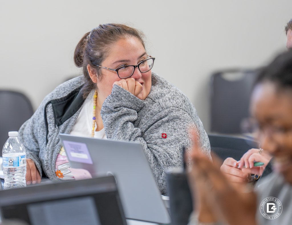A female educator in a gray jacket with glasses smiles while listening to a colleague during training. A laptop and water bottle sit in front of her.
