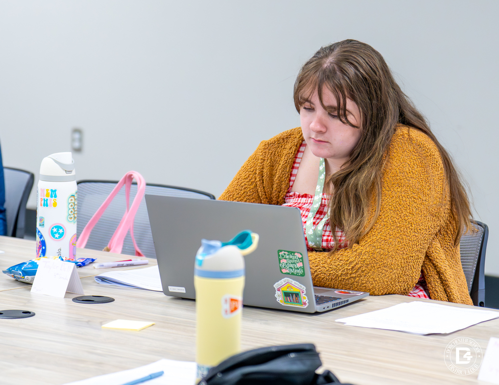 A female educator wearing a brown sweater focuses on her laptop while participating in training, with a water bottle and papers nearby.
