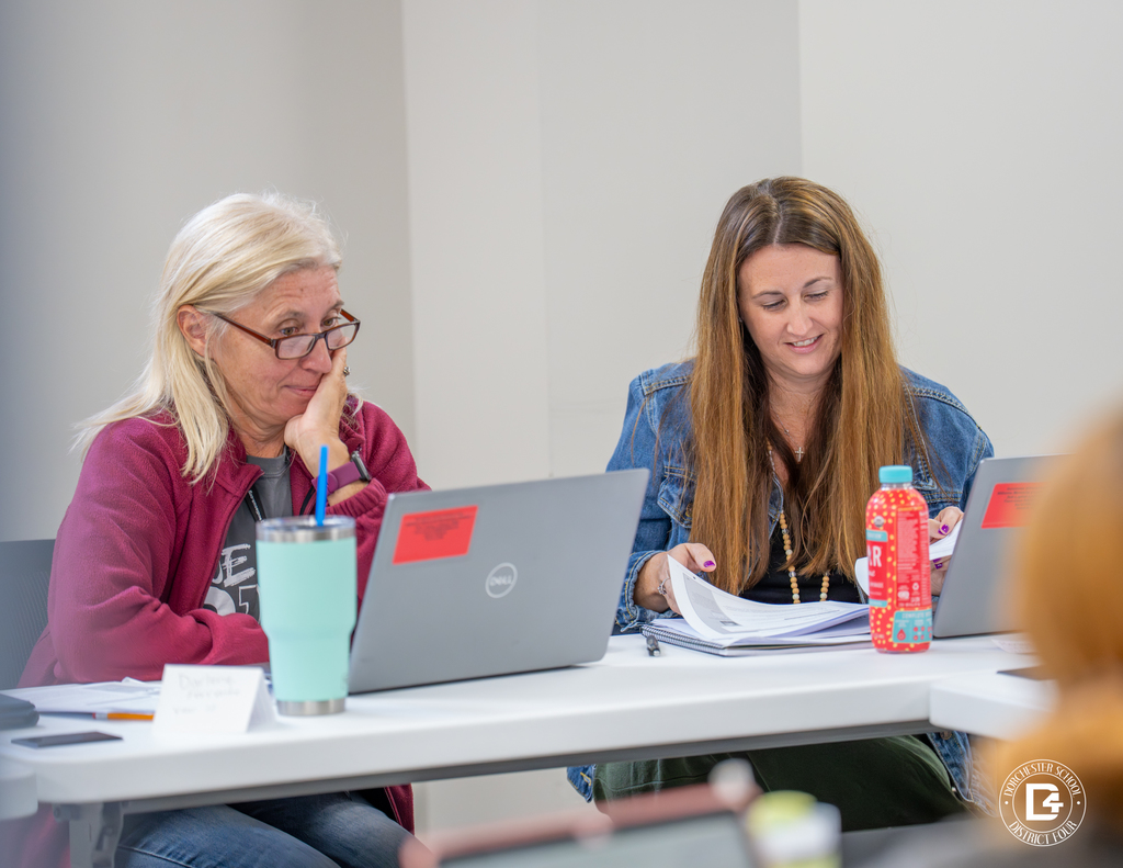 Two female educators, one with glasses and one with long hair, review training materials together while working on laptops.