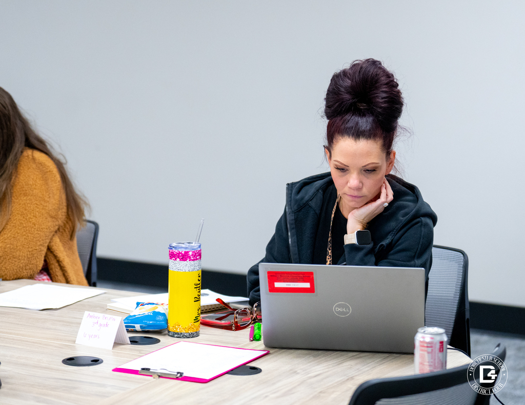A female educator with a high bun hairstyle works intently on her laptop during training. A drink cup and notes are on the table in front of her.