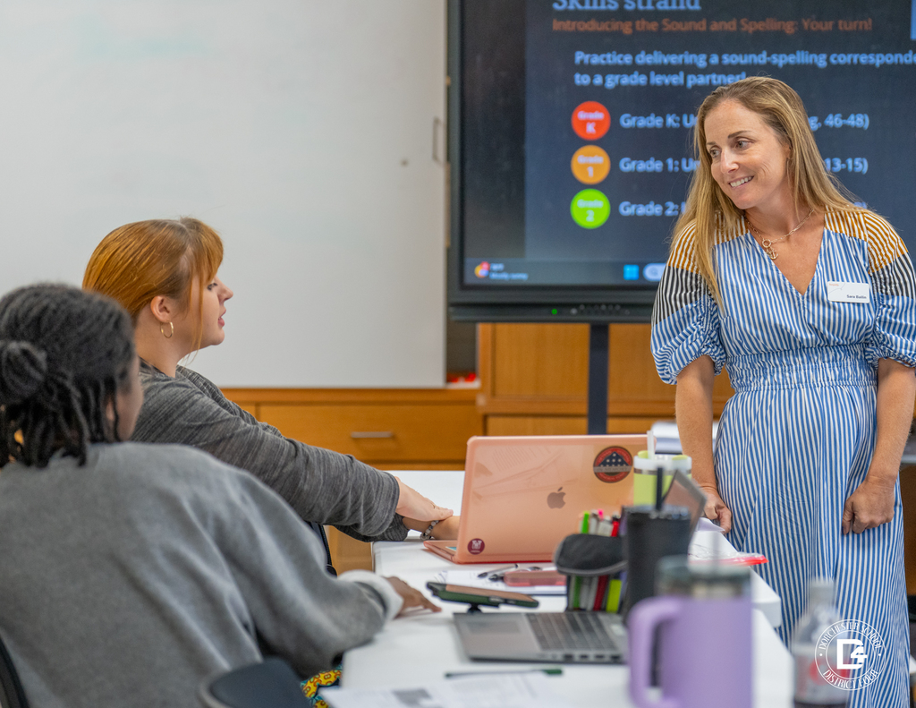 A female facilitator in a blue-striped dress stands and smiles while interacting with educators seated at a table, with a presentation screen in the background.