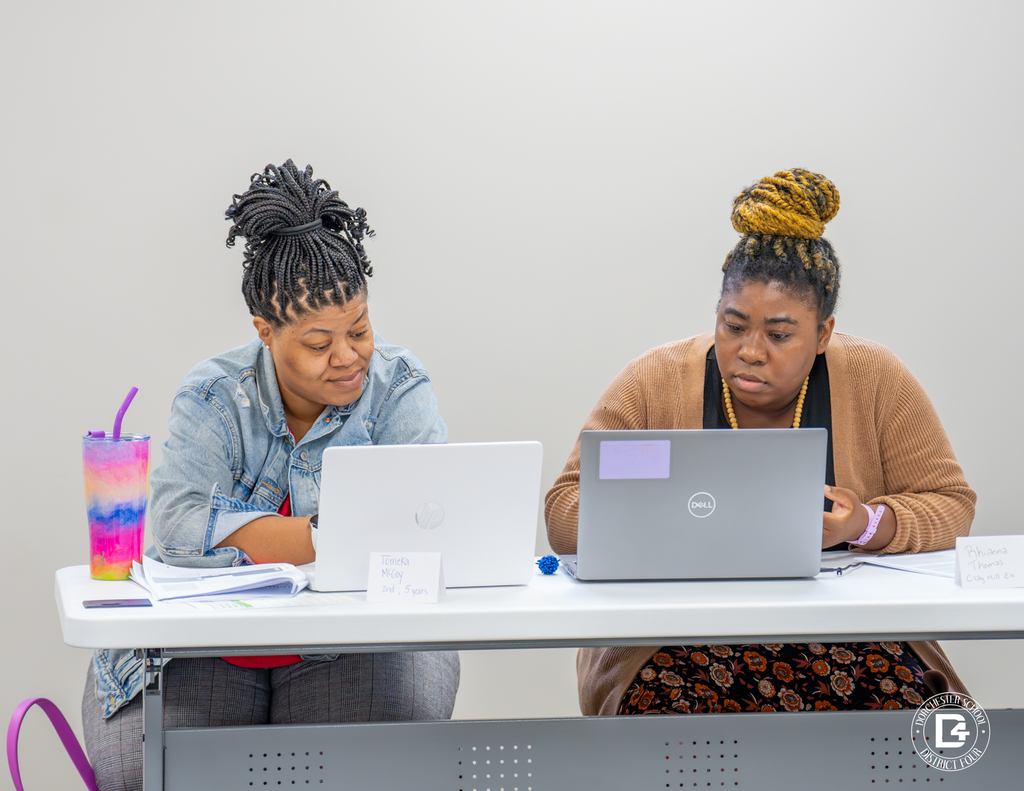 Two female educators sit side by side, working on laptops and discussing notes during training. Both have drinks and materials on the table.