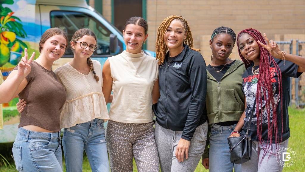 Six Woodland High School students smile and pose together outdoors, some flashing peace signs. A Kona Ice truck with a tropical parrot is in the background.