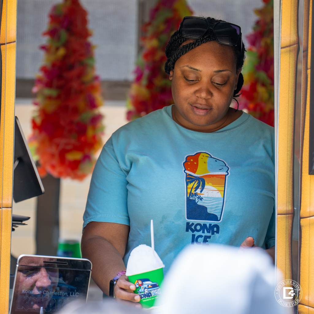 A Kona Ice worker serves students from a brightly decorated tropical-themed Kona Ice truck as a line of students waits.