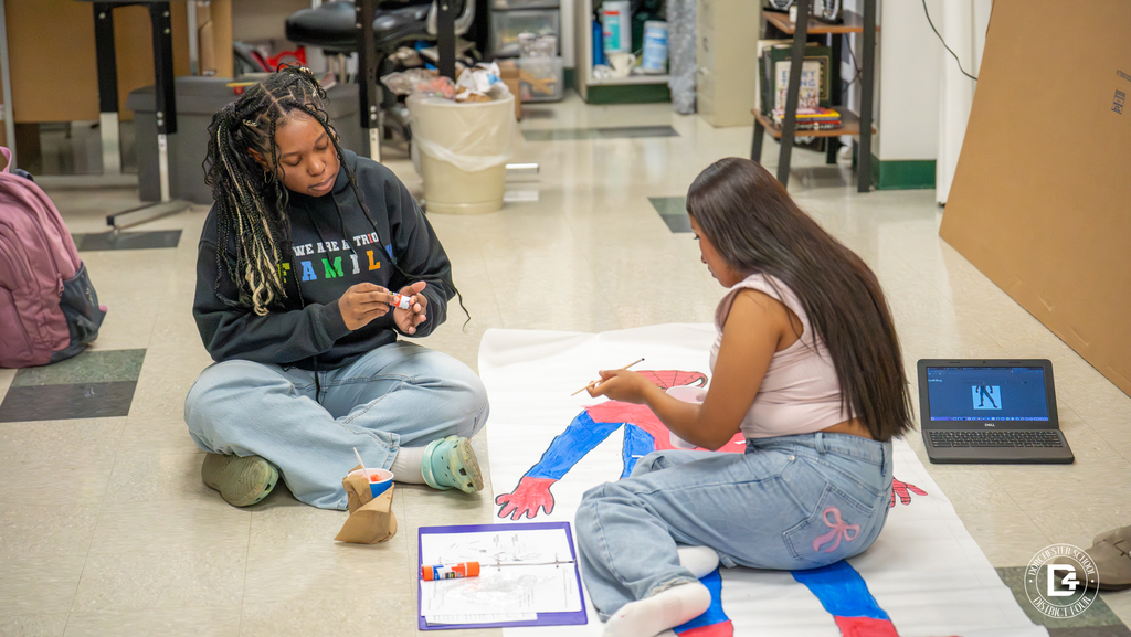 Two students collaborate on painting a Spider-Man poster laid out on the floor. A laptop nearby shows a reference image.