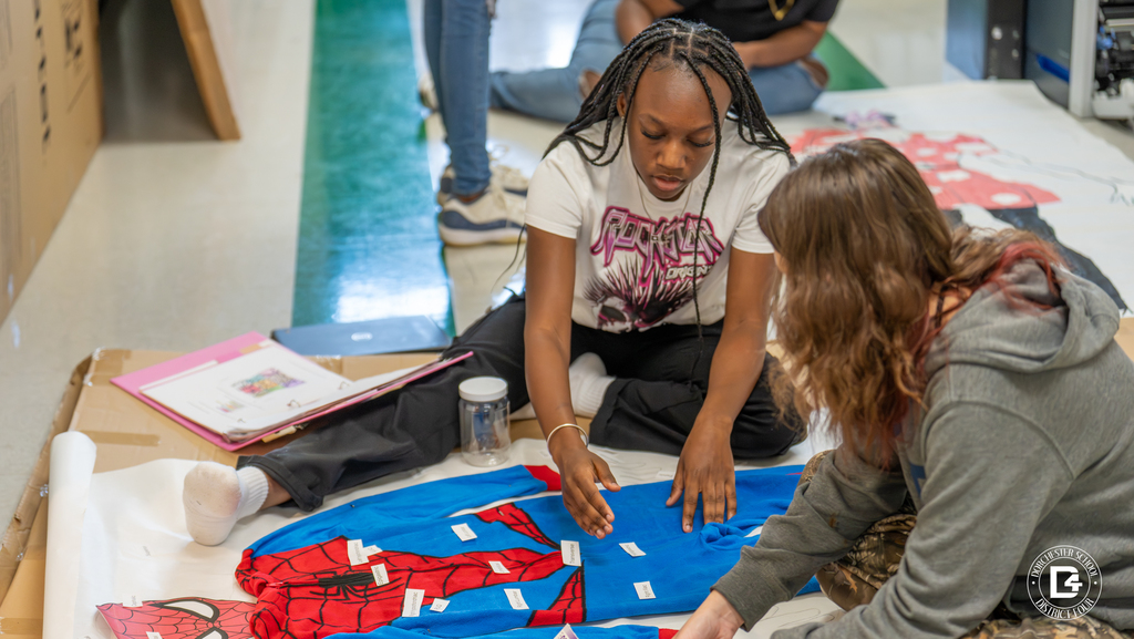 Two students sit on the floor working on a Spider-Man poster. One student gestures toward the poster while the other leans forward to place labels.