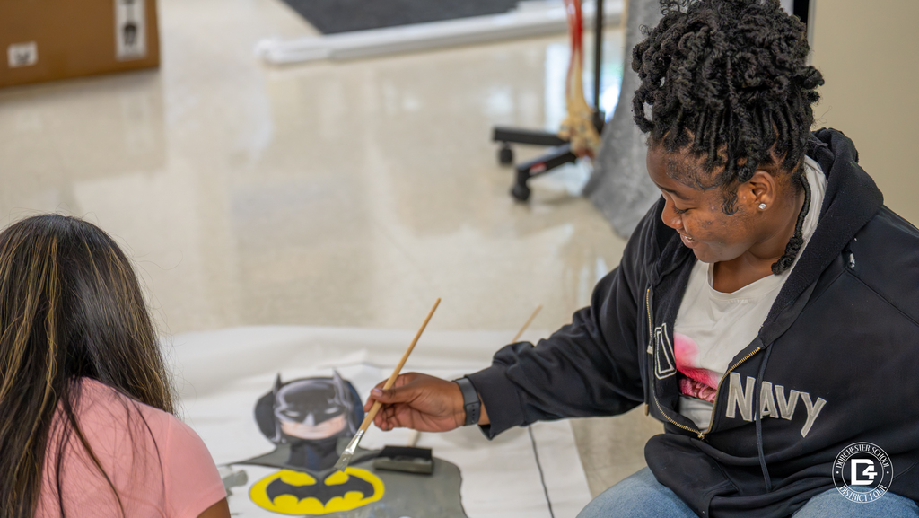 A student paints a large Batman poster on the floor, focusing on the character’s chest emblem, while smiling at their work. Another student sits nearby.