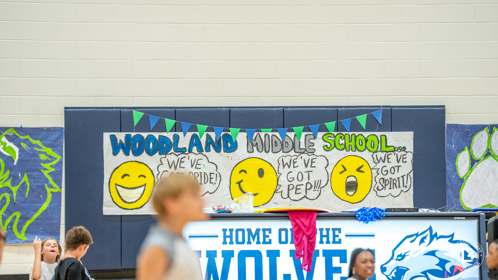A large painted banner in the gym reads “Woodland Middle School... We’ve Got Pride! We’ve Got Pep! We’ve Got Spirit!” decorated with smiling emoji faces and blue-and-green designs.