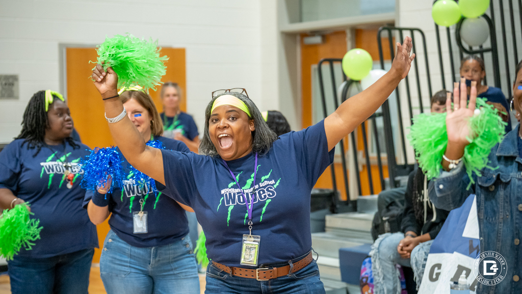 A Woodland Middle staff member waves green pom-poms in the air with a big smile while celebrating alongside other staff and students.