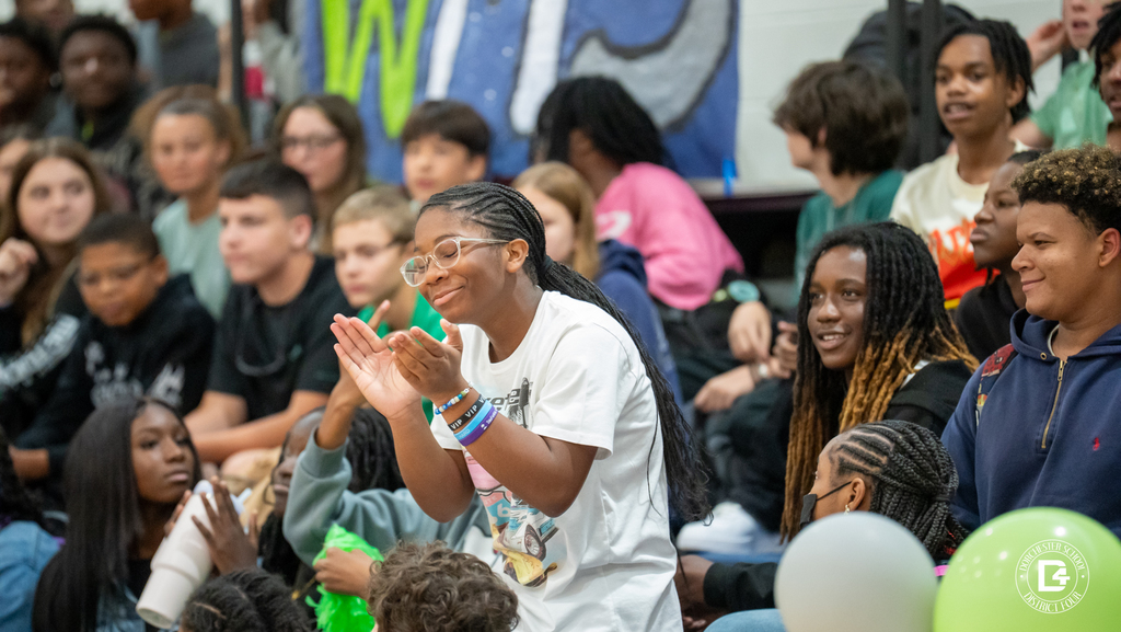 A student in a white shirt stands up clapping joyfully while surrounded by classmates who cheer and smile during the pep rally