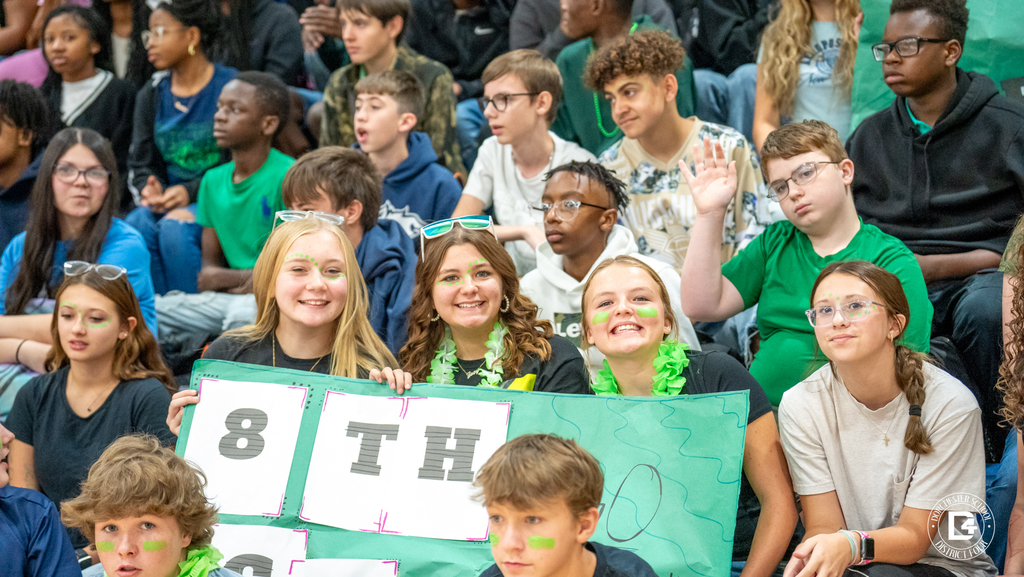 A group of eighth-grade students wearing green spirit gear and face paint smile and hold up a decorated sign that reads “8th.”