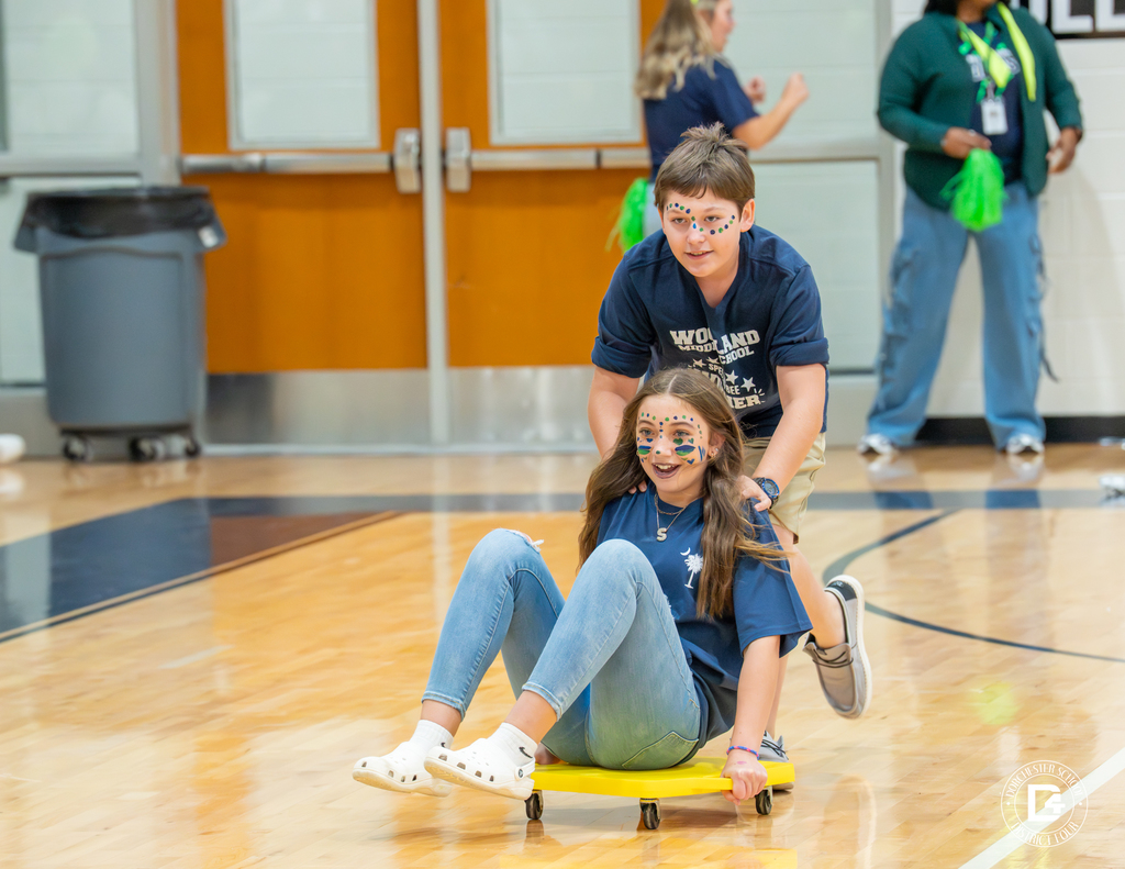 Two students participate in a scooter game in the gym, one sitting on a yellow scooter board while the other pushes them across the floor, both wearing face paint and smiling.