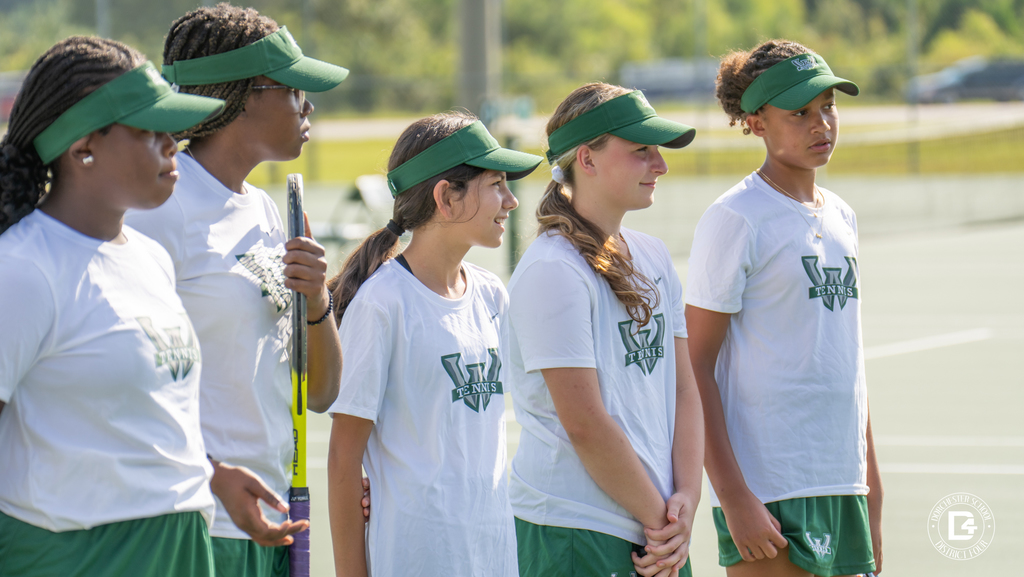 Woodland High School girls’ tennis players stand in a row on the court, wearing matching green visors and white shirts with the Woodland “W” logo.