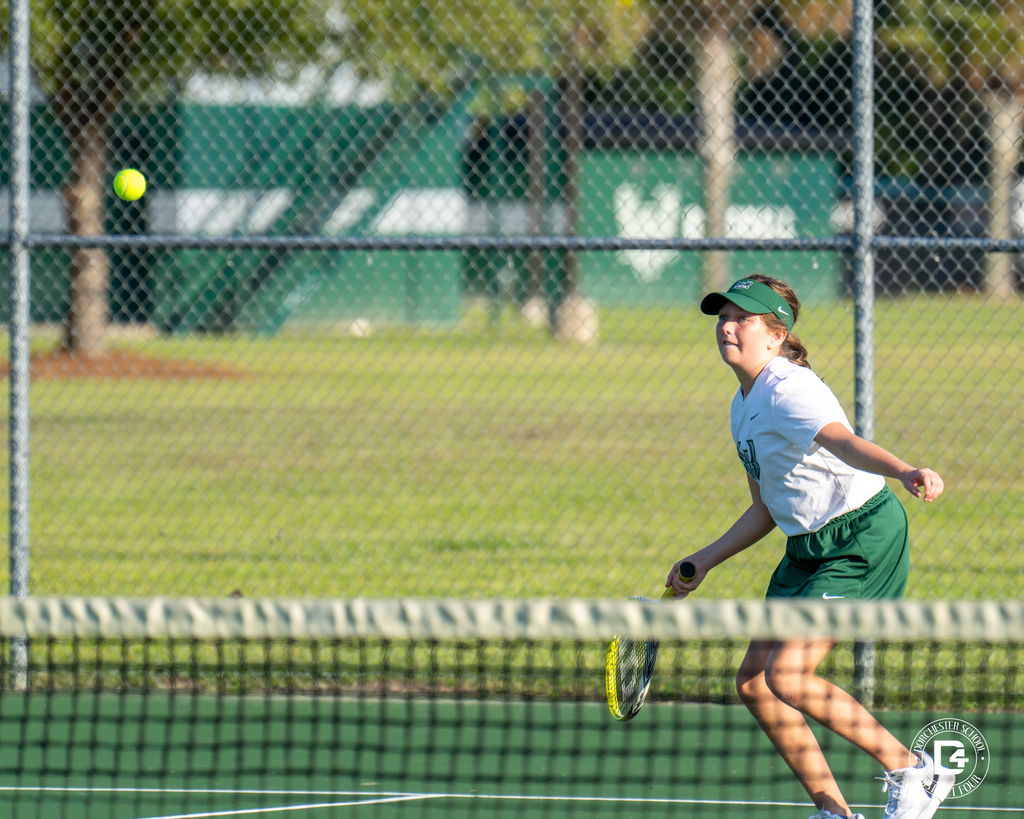 A Woodland High School tennis player leans forward, preparing to hit a shot as the ball approaches the net.
