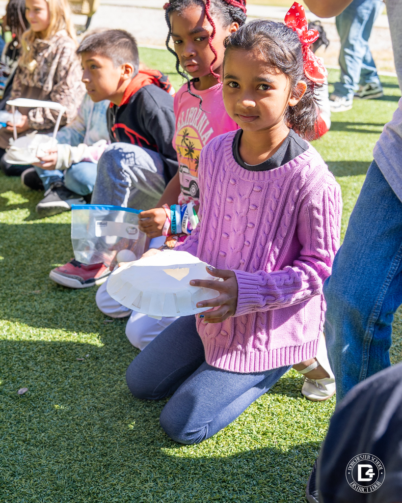 A girl in a pink sweater kneels on the grass holding her egg drop design made from a paper plate, while other students sit around her with their projects.