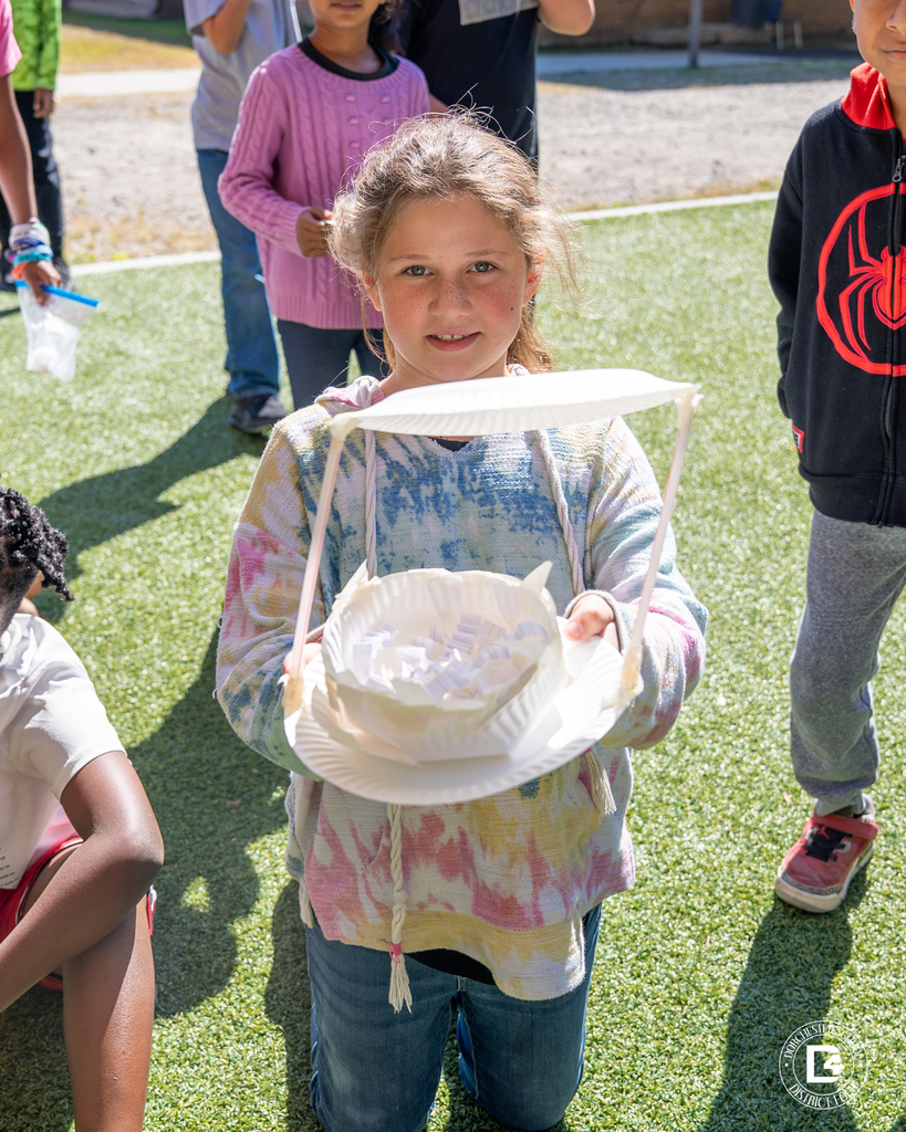 A student in a tie-dye hoodie kneels on the grass, proudly holding her egg drop device built from paper plates, tape, and paper strips.