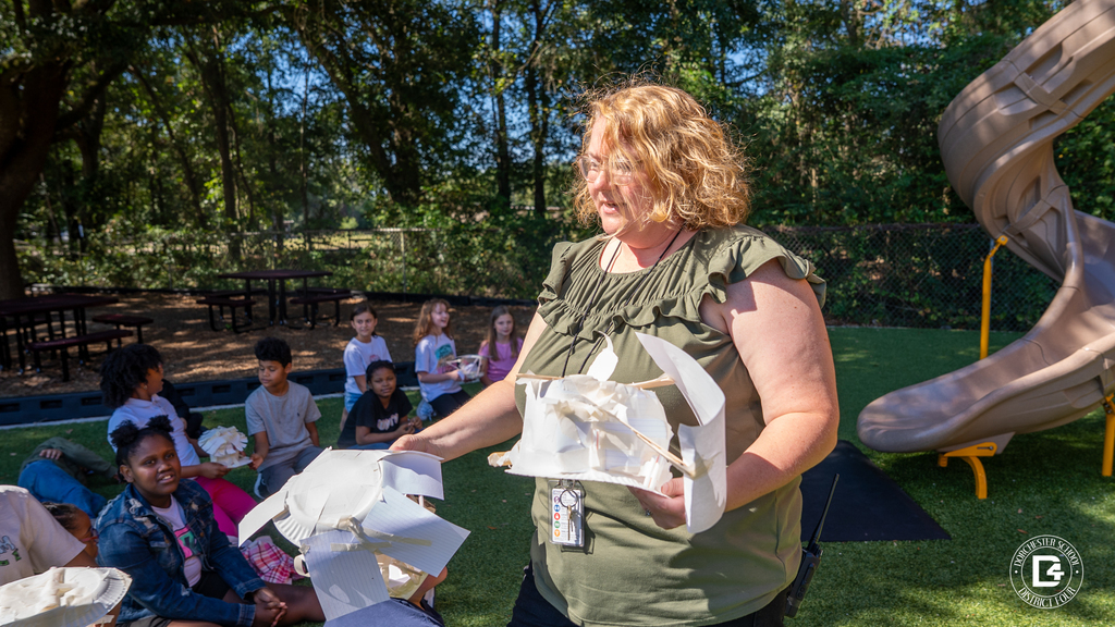 A teacher holds up several student-made egg drop contraptions while addressing a group of children seated outside near the playground.