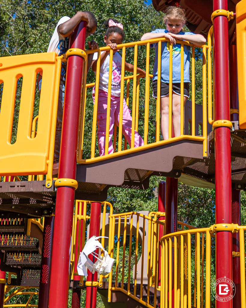 Students stand on a tall playground structure as one of them releases an egg drop contraption, which is captured midair falling to the ground.