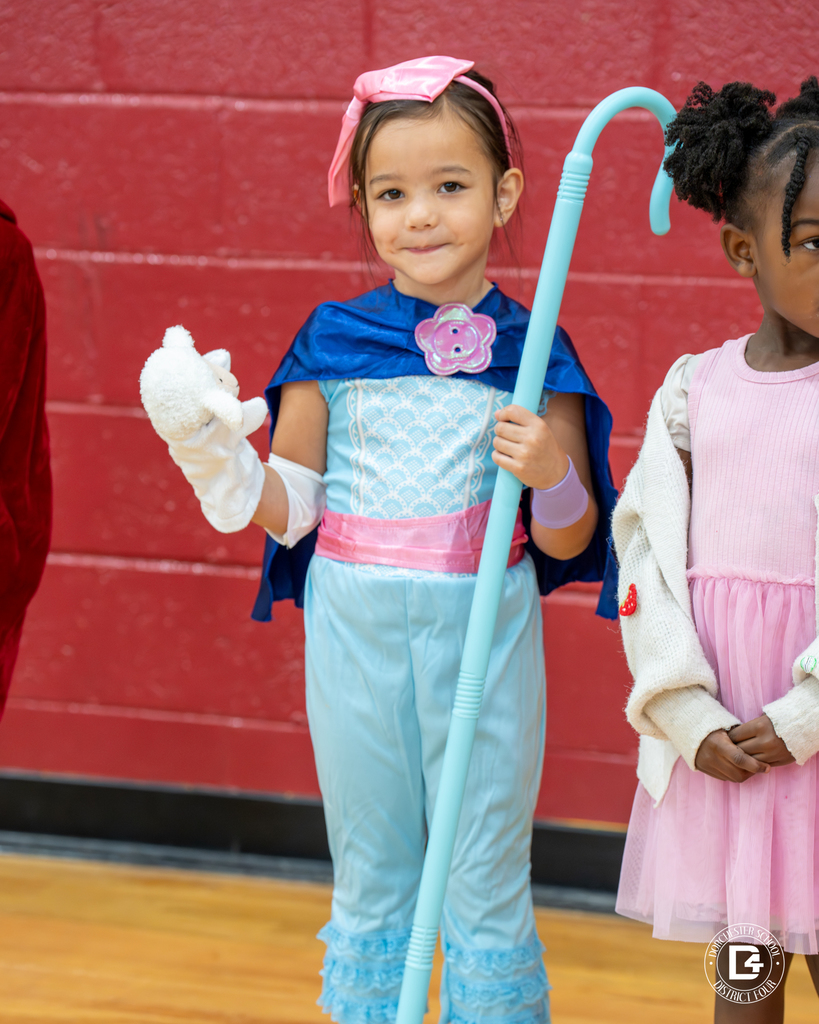 A girl dressed as Little Bo Peep holds a small lamb plush and shepherd’s staff, smiling at the camera.