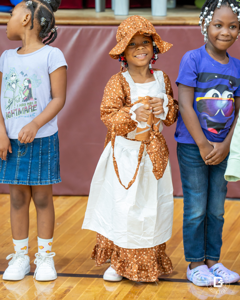 A girl dressed in a brown bonnet and dress with a white apron smiles brightly, standing between classmates.