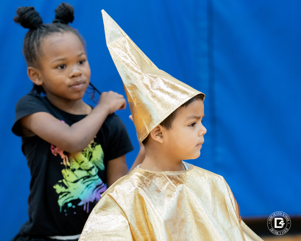 A boy in a shiny gold cone hat and matching robe stands in profile next to a girl in a black T-shirt.