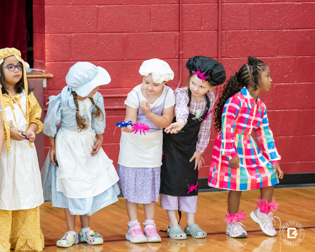 Five girls in pioneer and nursery rhyme costumes line up, some holding toy spiders and wearing bonnets or aprons.