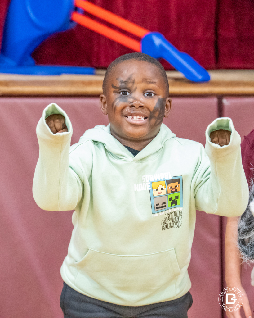 A boy with black face paint grins and raises his fists while wearing a light green Minecraft hoodie.