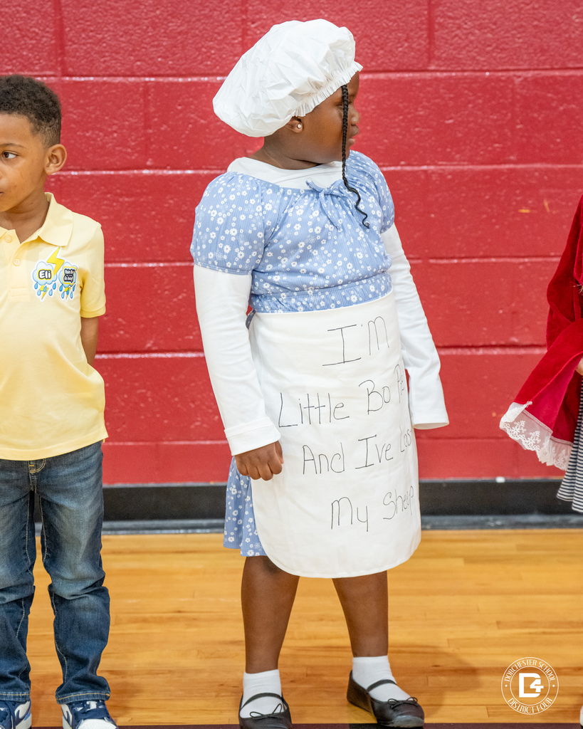 A girl dressed as Little Bo Peep wears a blue dress, white bonnet, and apron that reads “I’ve lost my sheep.”