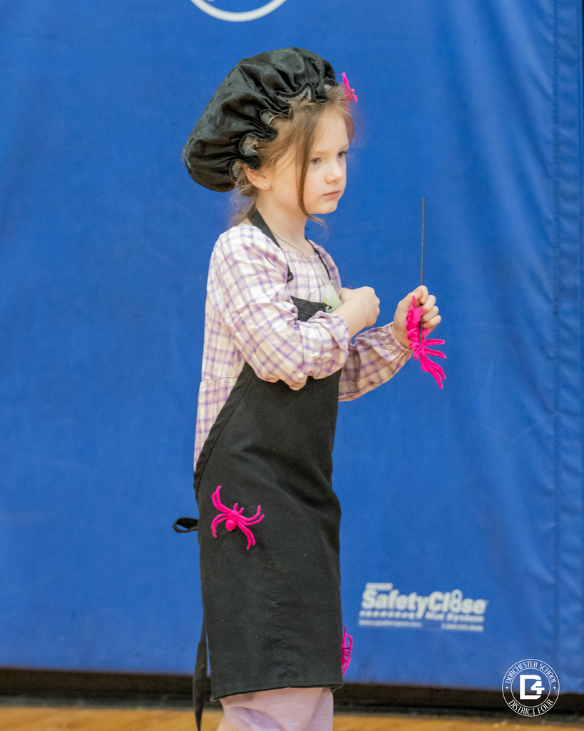 A girl dressed as Little Miss Muffet wears a black bonnet and apron decorated with pink toy spiders.