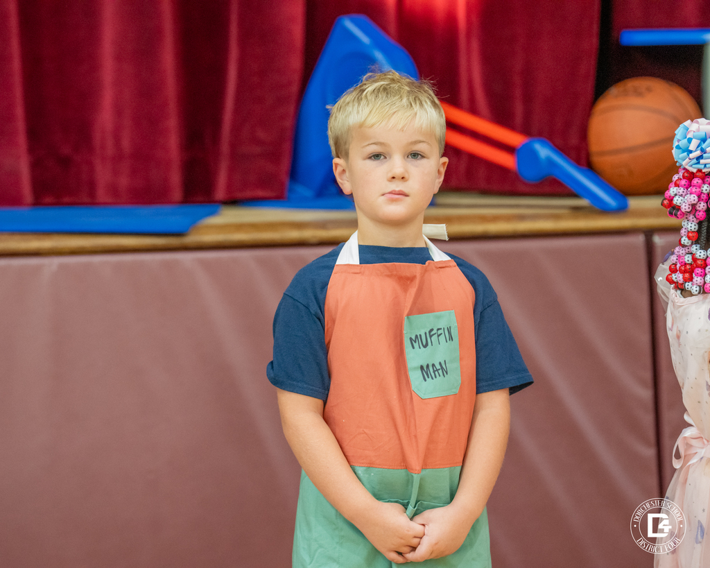 A boy dressed as the Muffin Man stands in front of a stage, wearing an apron labeled “Muffin Man.”