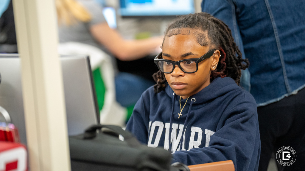 A student in a navy blue “Howard” hoodie and glasses works at a computer, concentrating on the screen. A backpack and water bottle are visible on the desk nearby.