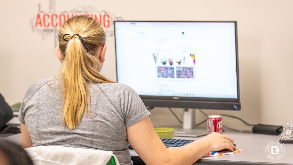 A student with blonde hair in a ponytail sits at a desk, working on a computer. The screen displays a page layout with photos and text, while a soda can and notebook sit nearby.