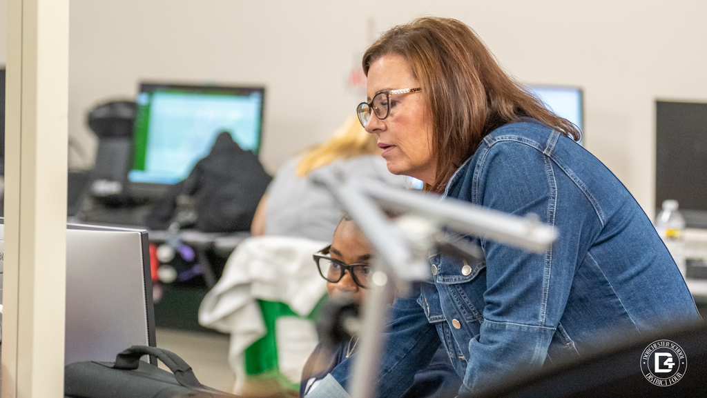 A teacher in a denim jacket leans over to assist a student at a computer. The student, wearing glasses, looks at the screen while the teacher provides guidance.