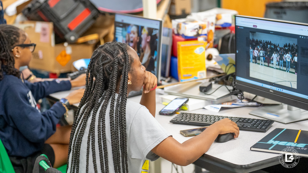 Two students sit side by side at separate computers, reviewing photos of a school event on their screens. One student with long braids uses the mouse while the other looks on attentively.